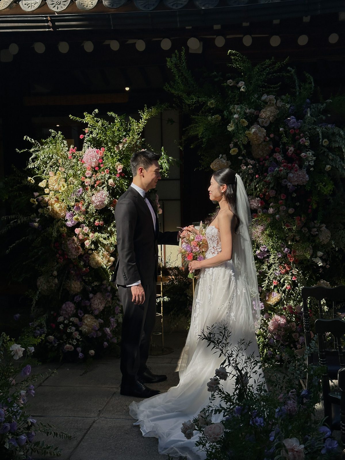 A bride and groom stand facing each other in front of a floral backdrop, dressed in wedding attire, outdoors in natural light.