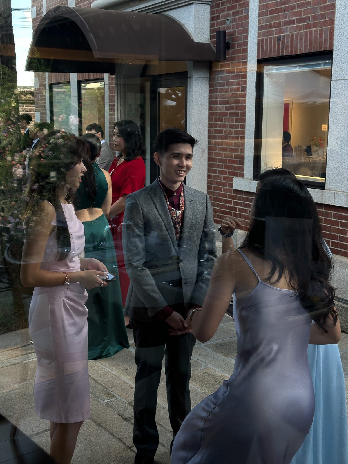A group of people dressed formally talk outside a building with brick walls and large windows, perhaps discussing what is a wedding content creator. One man in a gray suit and two women in dresses are in the foreground.