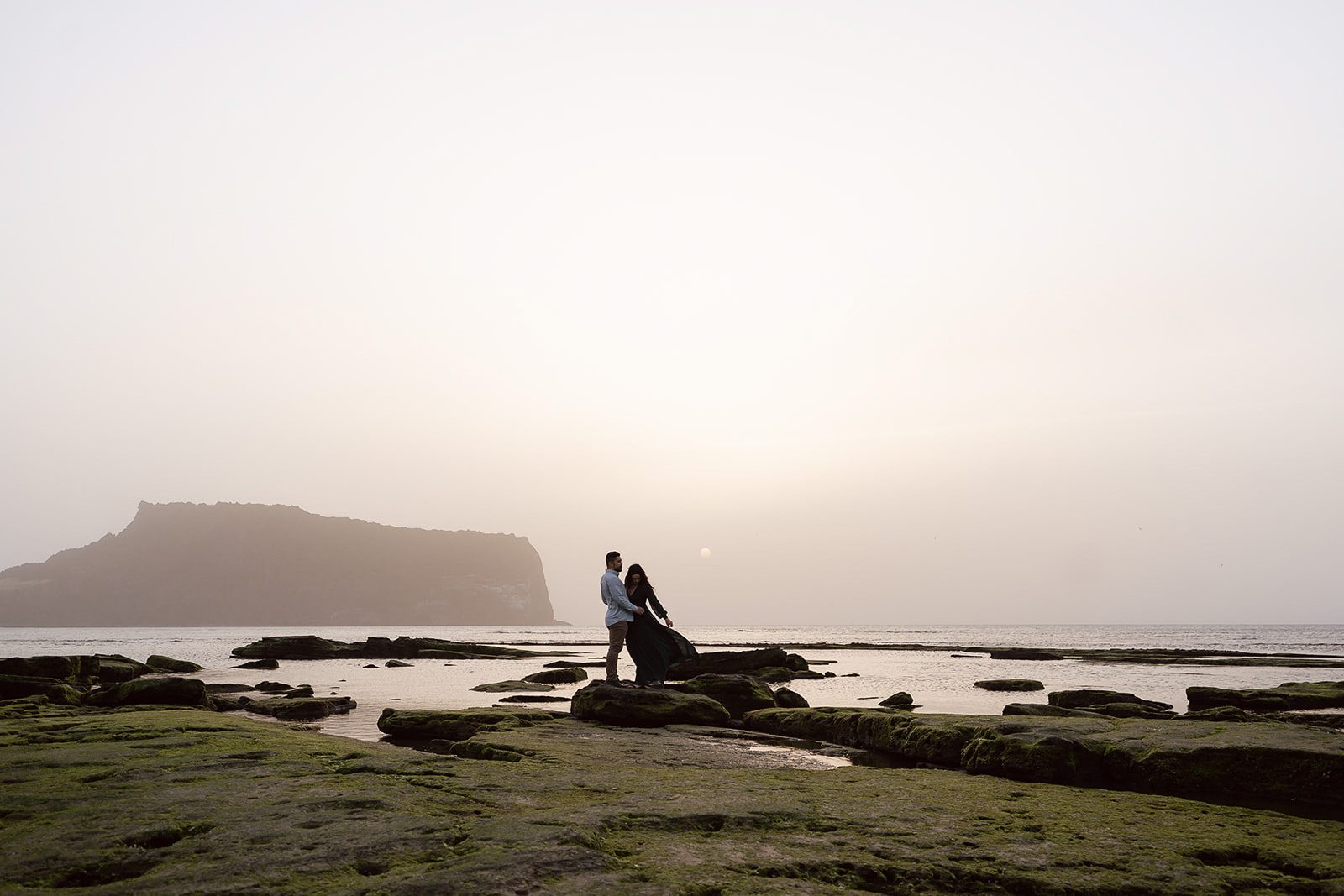 A couple stands on mossy rocks at a coastal area during sunset, with a large rocky island visible in the distance under a hazy sky.