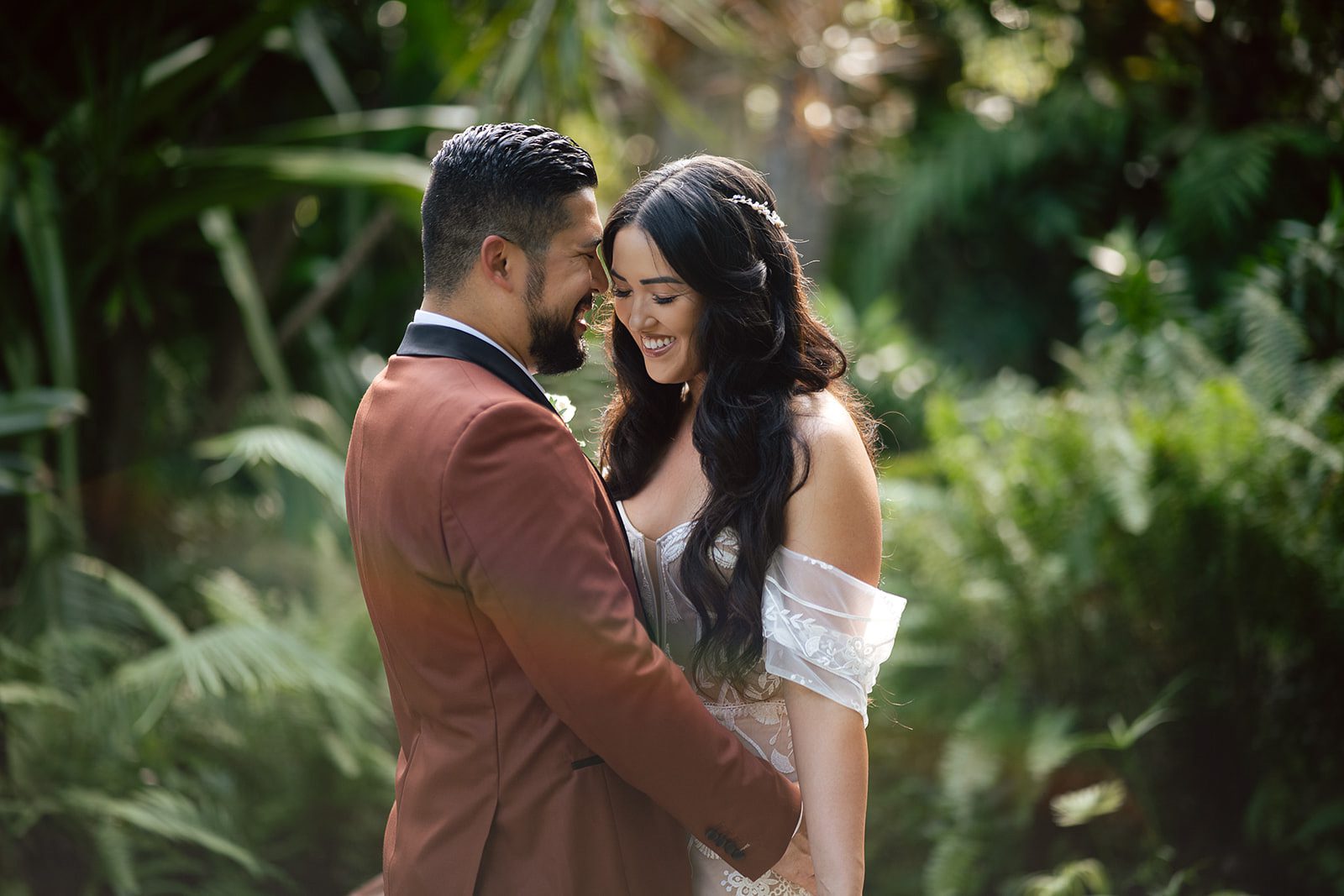 A bride and groom stand close together outdoors, smiling at each other amidst lush green foliage, capturing the romance of a Bali destination wedding.