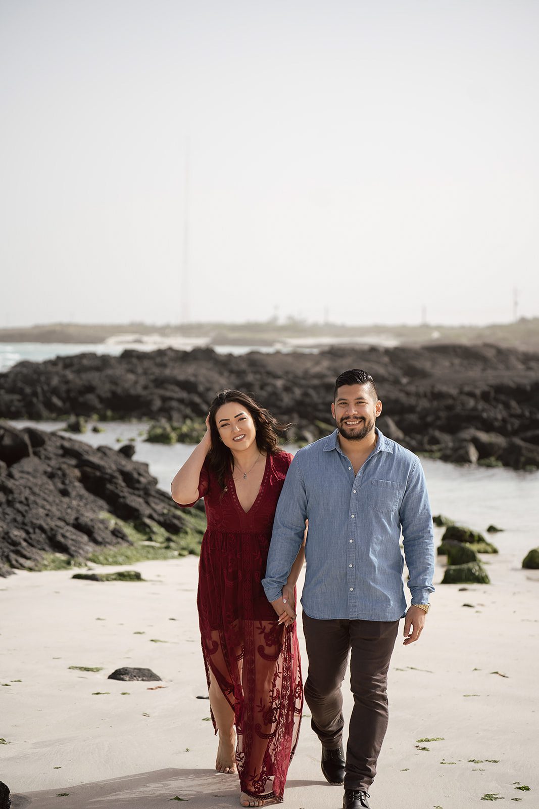 A woman in a red dress and a man in a blue shirt walk together on a rocky beach taking pre-wedding photos before their Bali destination wedding. 