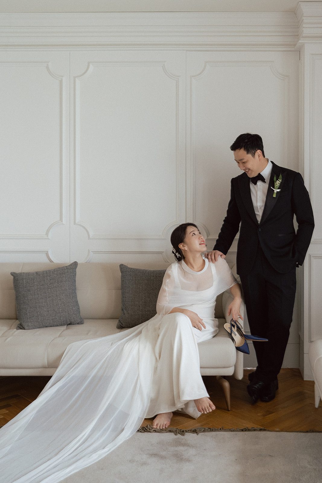 A bride in a white dress sits barefoot on a sofa, holding her shoes, while a groom in a black suit stands beside her; they look at each other and smile in this elegant Korean wedding photography scene.