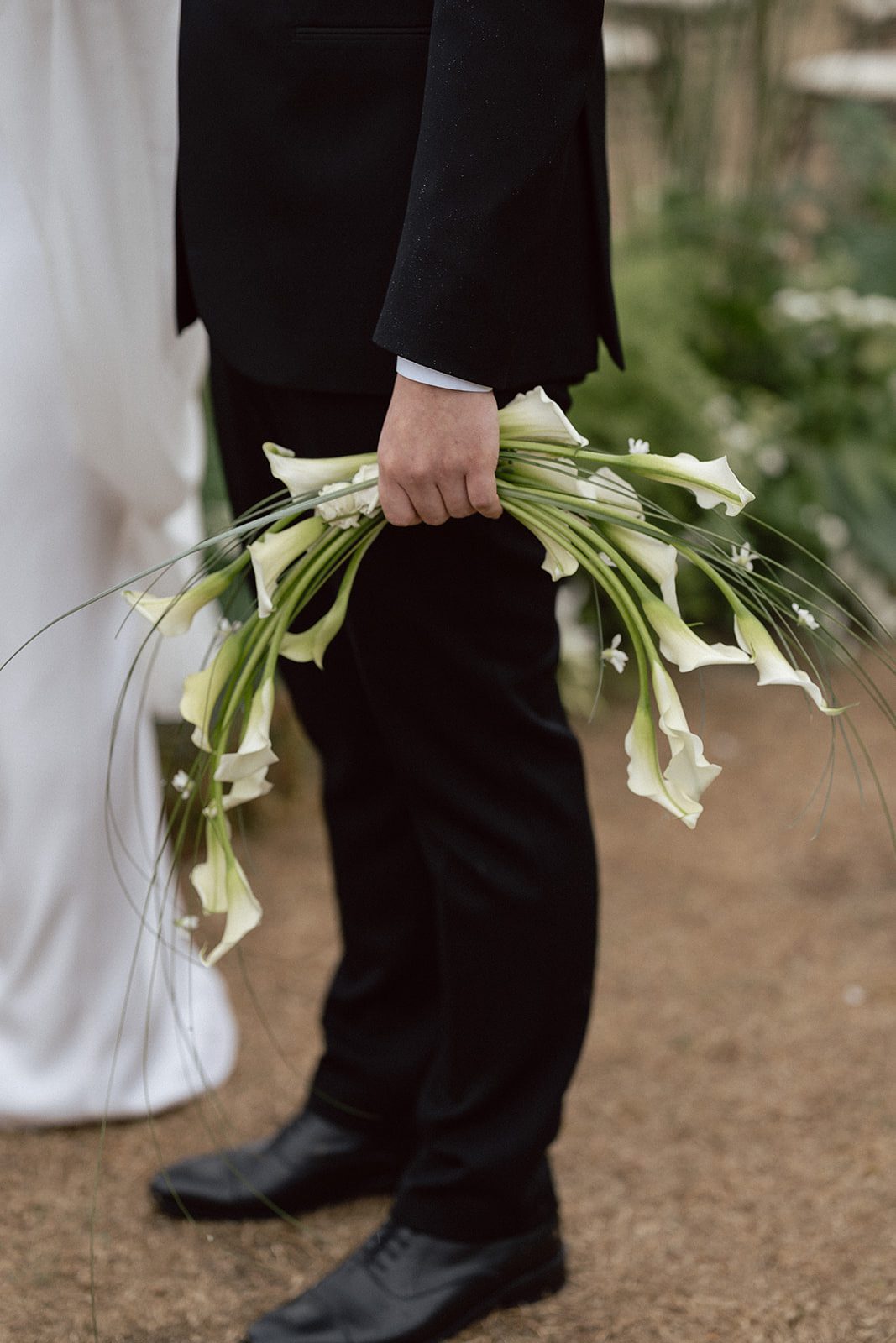 A person in a black suit holds a bouquet of white calla lilies, standing on a dirt pathway with greenery in the background—perfect for elegant wedding photography inspired by Korean style.