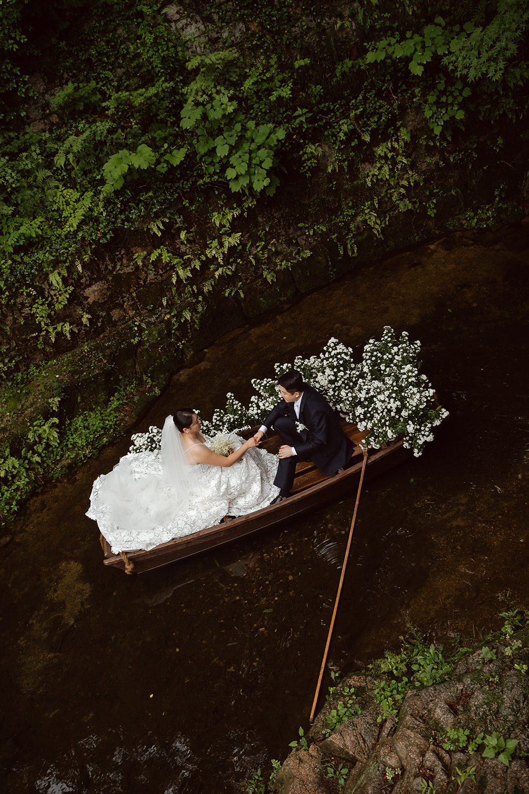 A bride and groom in wedding attire sit in a small flower-decorated boat on a narrow stream surrounded by lush greenery, capturing the elegance of Korean wedding photography.