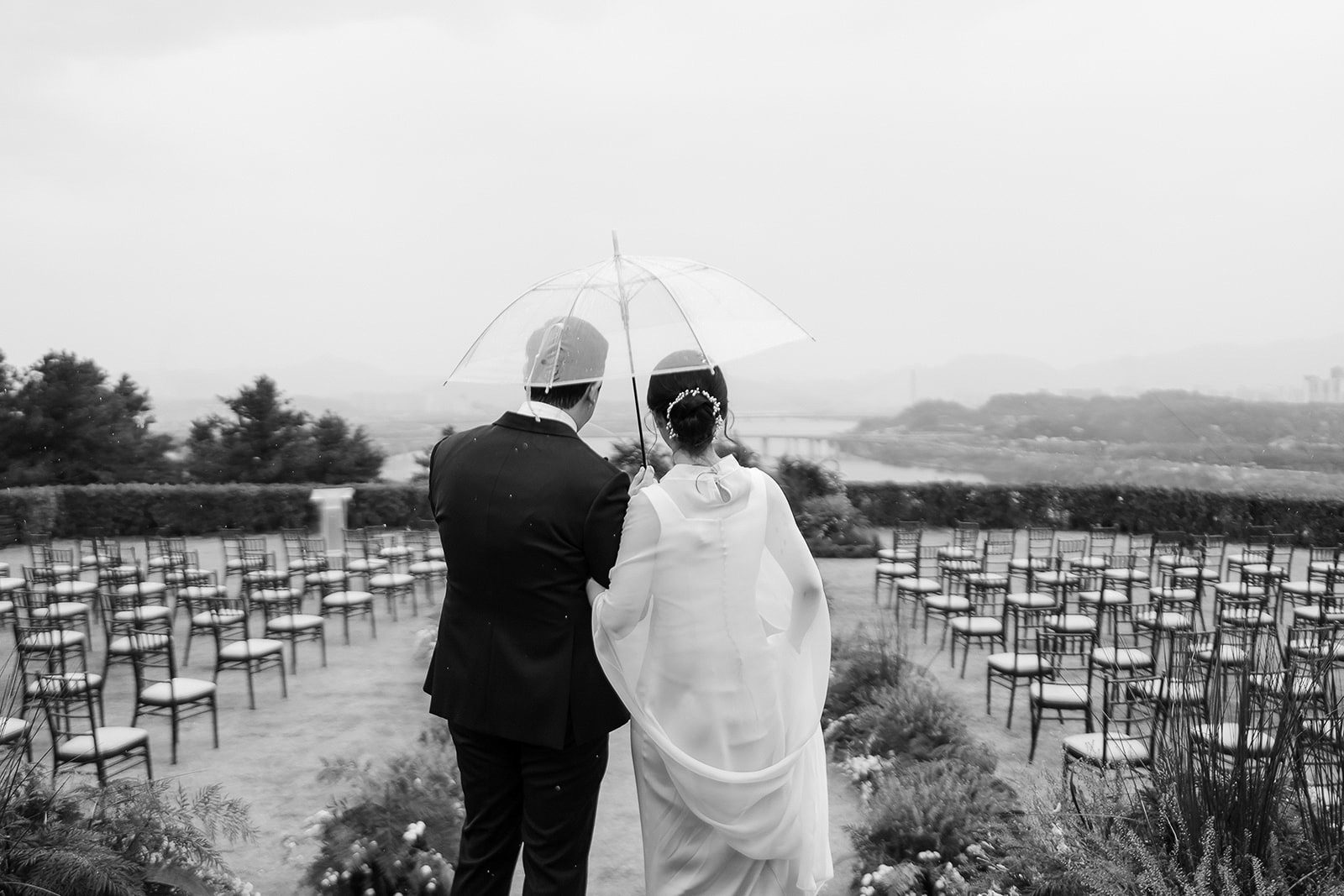 A couple in formal attire stands under a transparent umbrella, facing an empty outdoor wedding seating area on a cloudy day, capturing the timeless charm of Korean wedding photography.