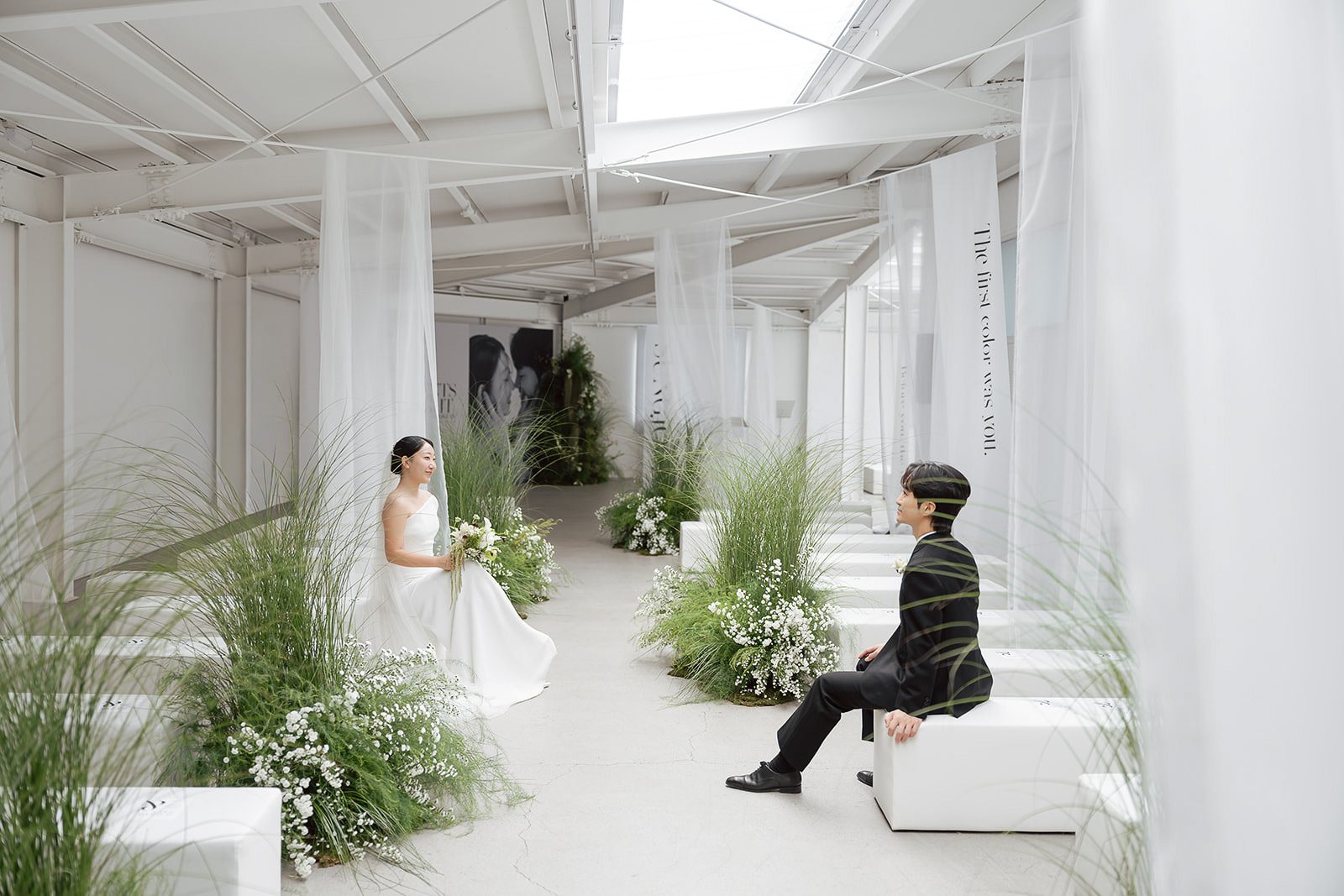 A bride and groom sit facing each other in a minimalist, white wedding venue decorated with sheer curtains, greenery, and white flowers—capturing the essence of elegant Korean wedding photography.