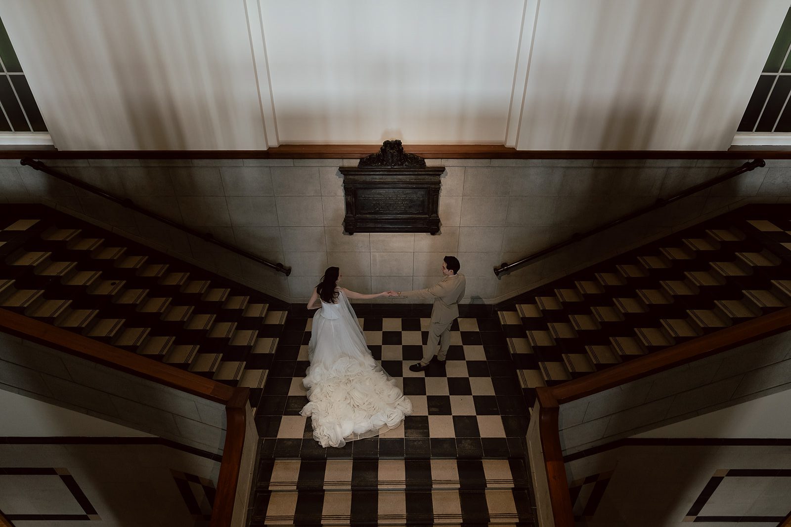 A bride and groom stand holding hands at the bottom of a grand staircase with black and white checkered flooring, viewed from above, part of their pre wedding shoot in Singapore.