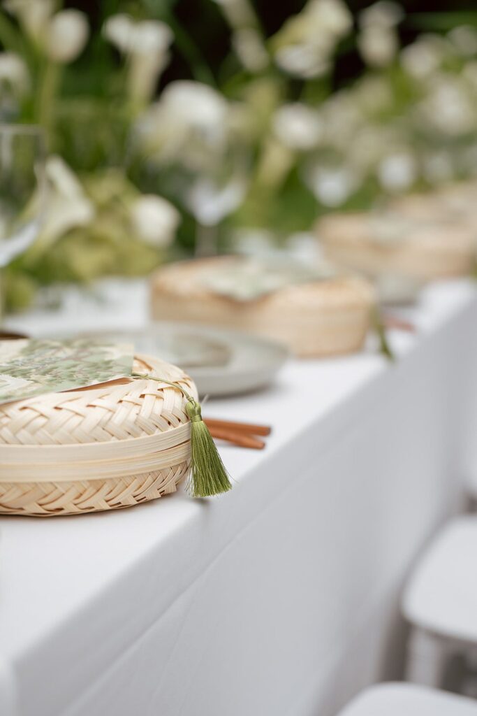 A close-up of a woven basket with a green tassel on a white tablecloth, set for an event with plates, utensils, and floral decorations in the background—capturing the refined details often highlighted in Korean wedding photography.