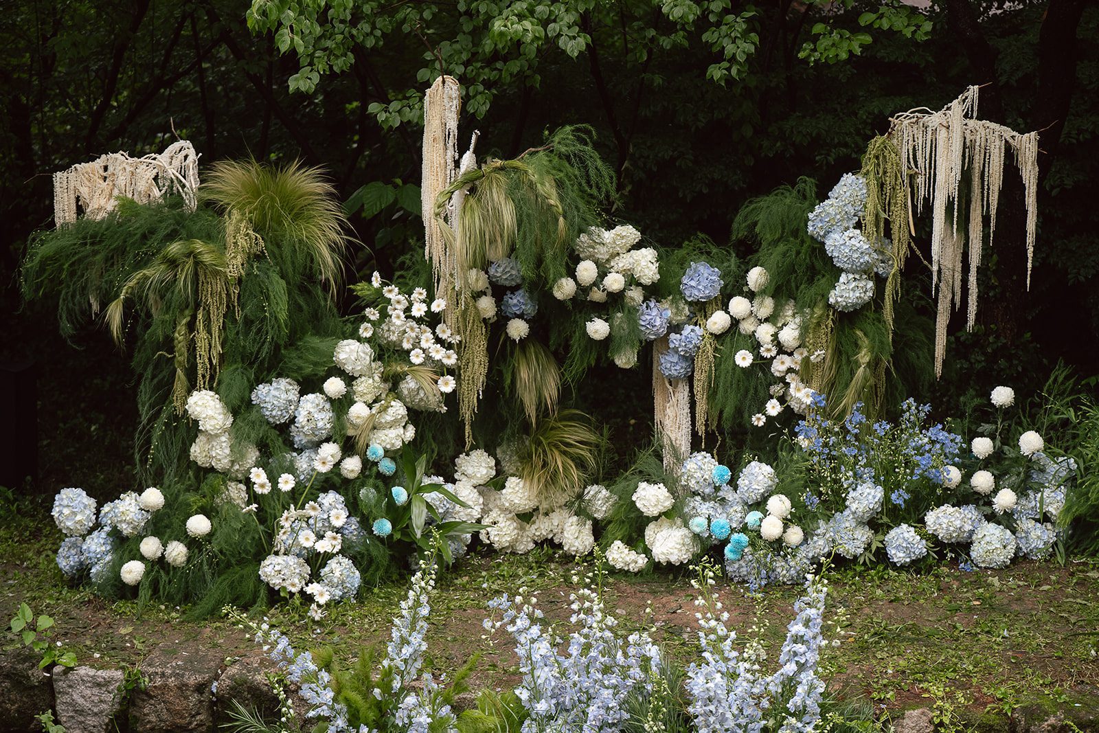 A lush outdoor floral arrangement, perfect for wedding photography, featuring white and blue hydrangeas, hanging greenery, and various white flowers set against a backdrop of trees and shrubs in elegant Korean style.