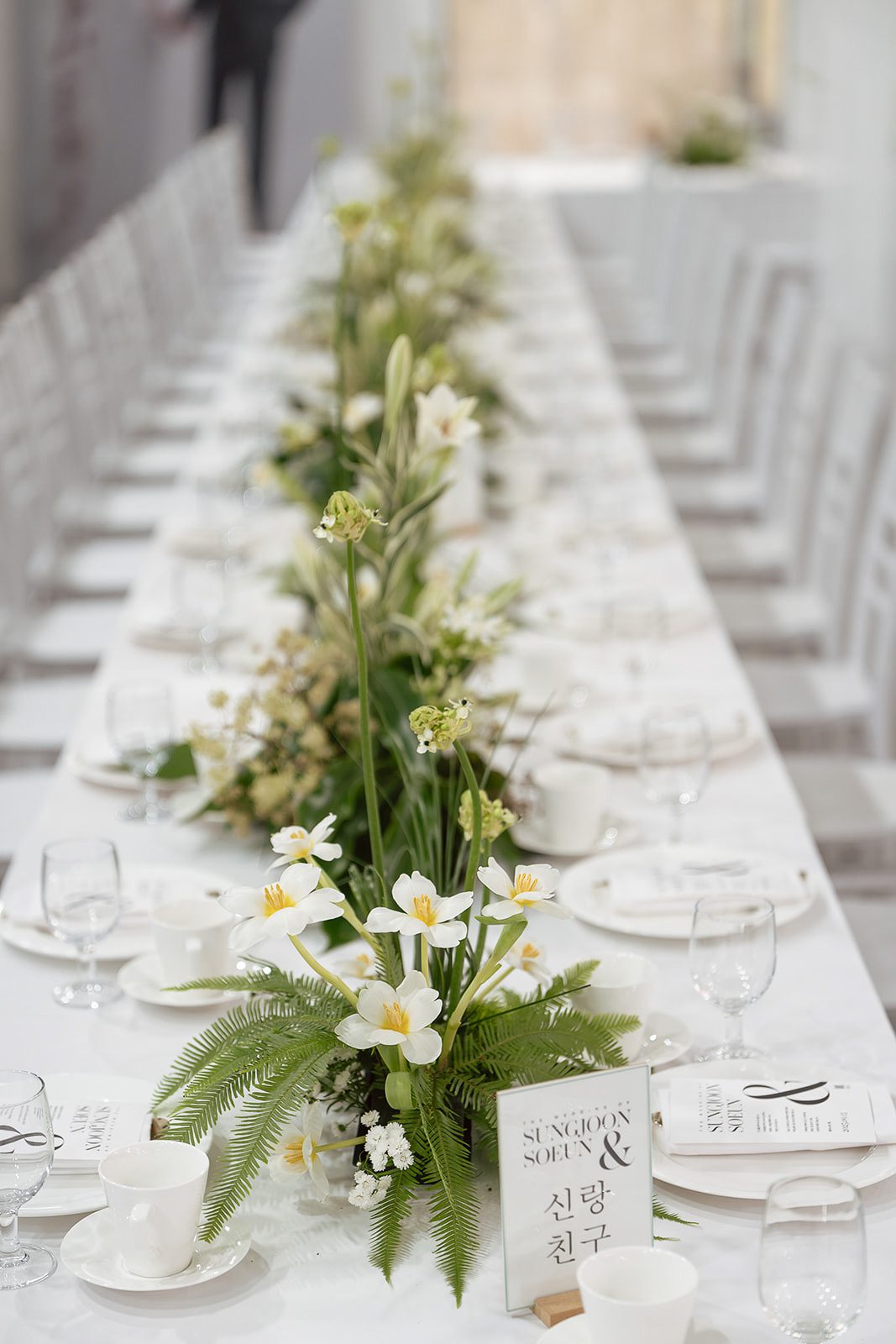 A long, white table set with white dishes, glassware, and floral centerpieces featuring white flowers and greenery.