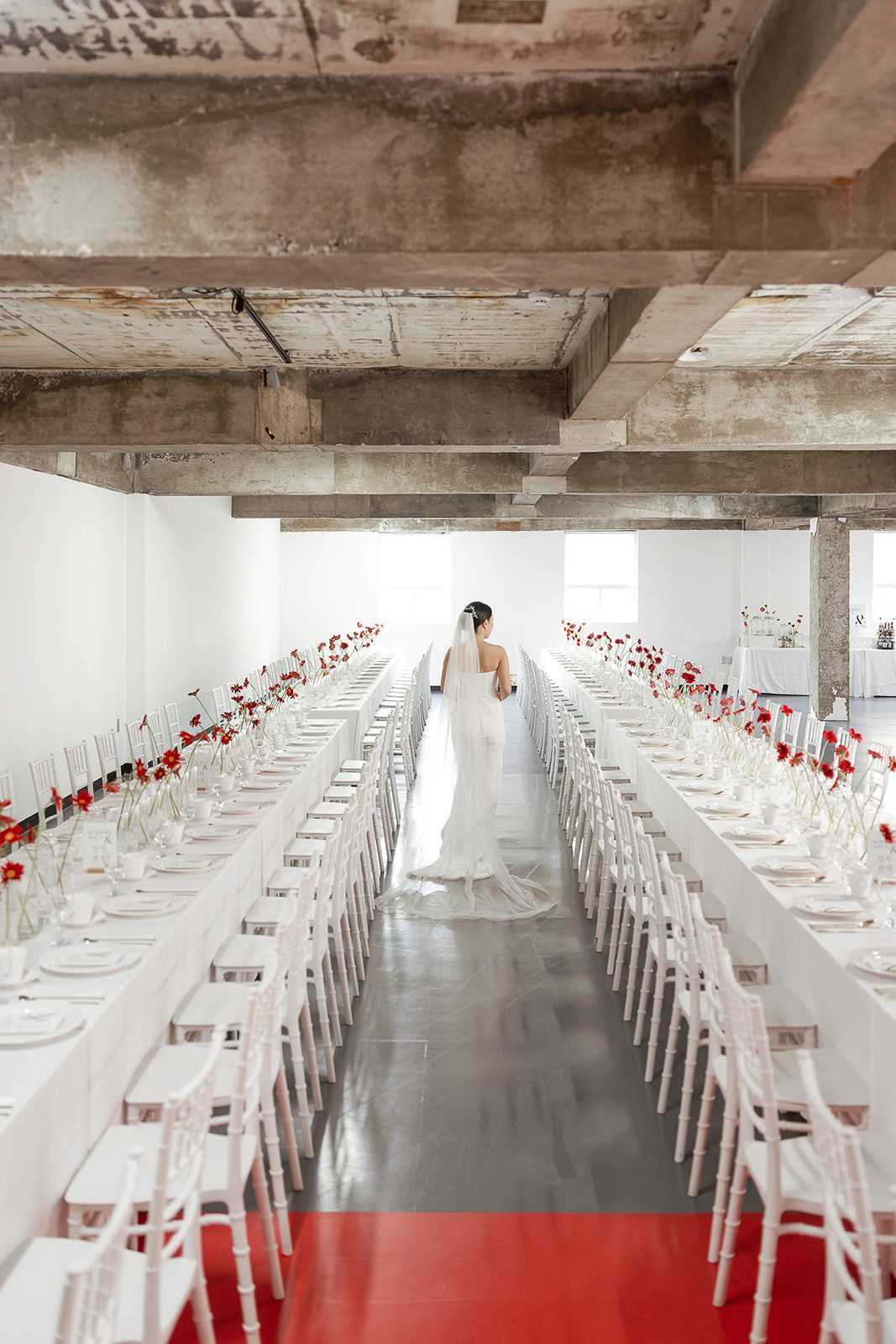 A bride in a white gown and veil stands in the center aisle between two long banquet tables set with red and white decor.