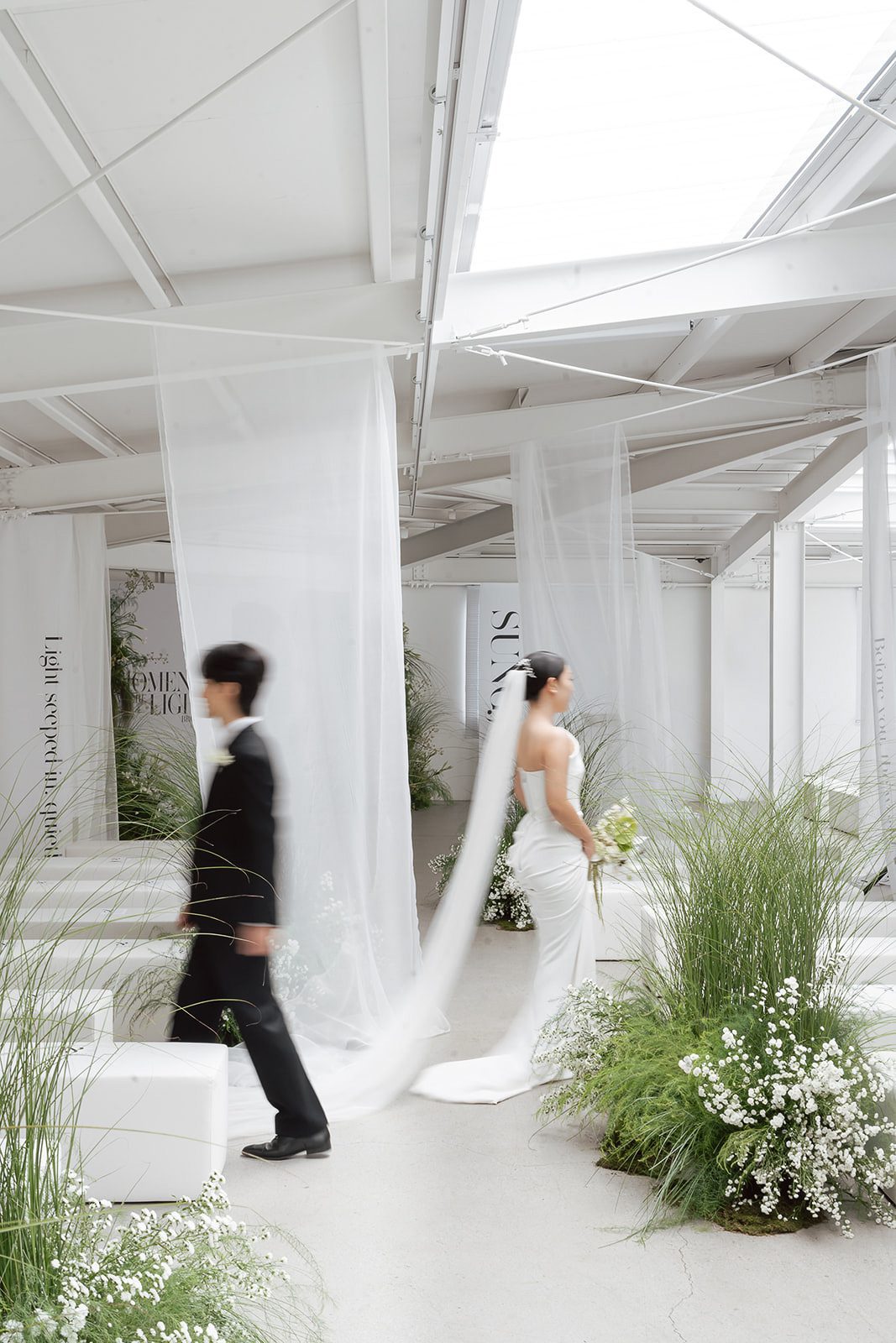 In this wedding photography scene, a Korean bride in a white gown and veil holds flowers as a man in a black suit walks by in a bright, modern venue adorned with greenery and sheer white drapes.