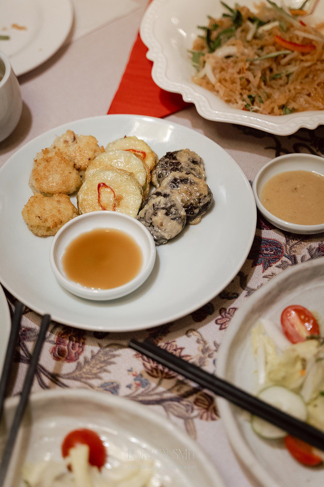 A white plate with assorted fried vegetables, a small bowl of dipping sauce, and other dishes like noodles and salad are artfully arranged on the table.