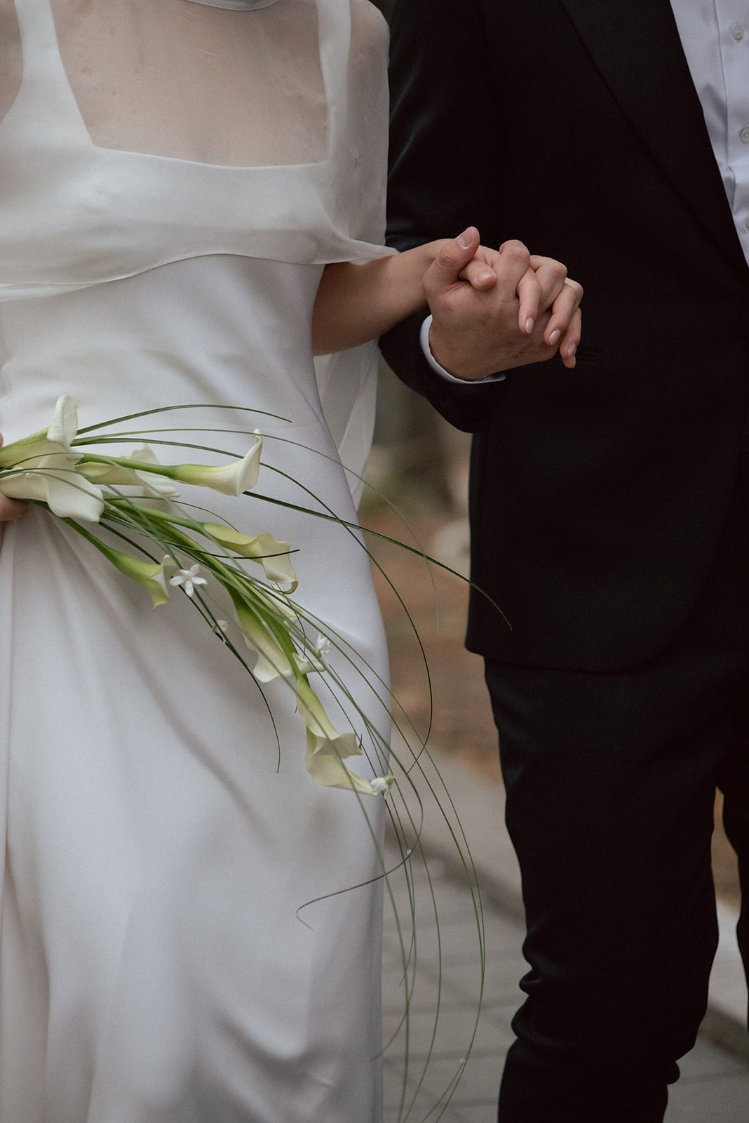 A bride in a white dress holds white calla lilies and the hand of a person in a black suit, as they walk together at their wedding in Korea.