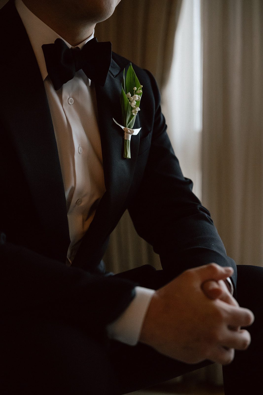 Man in a black tuxedo with a white shirt and black bow tie, wearing a boutonniere, sits with hands clasped in a dimly lit room, before the wedding ceremony