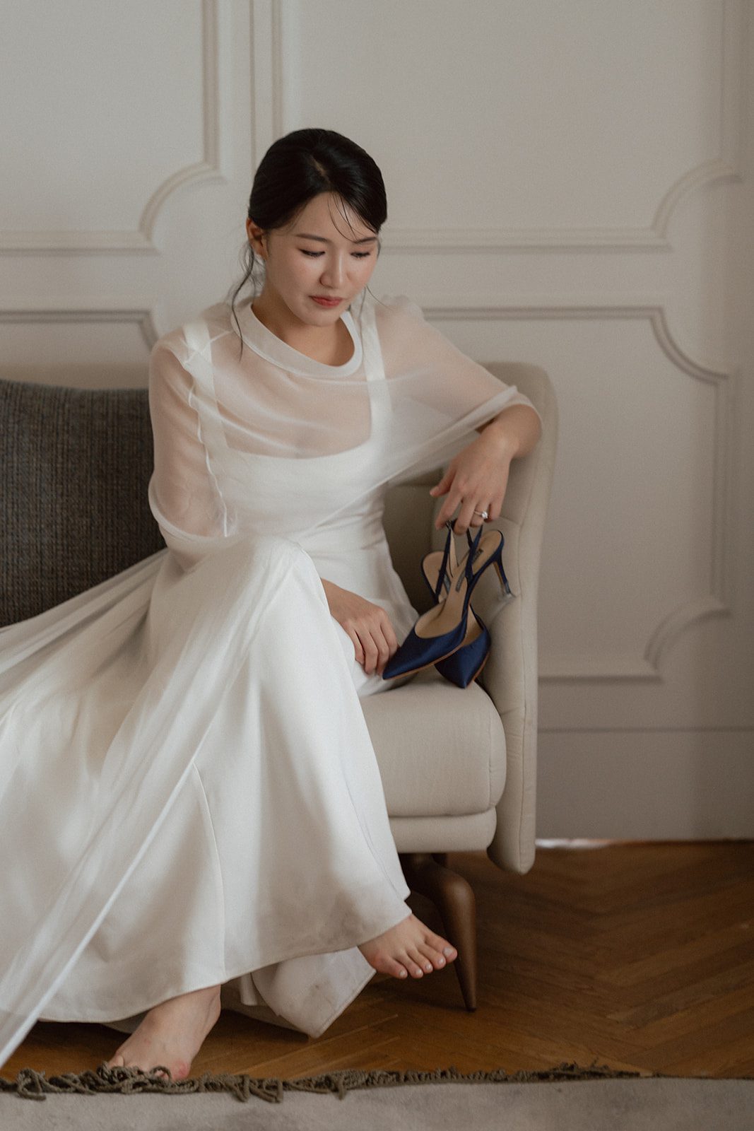 A woman in a white dress sits on a chair, holding navy high-heeled shoes in her hand with one bare foot resting on the floor, lost in thought about choosing wedding day venues in Korea.
