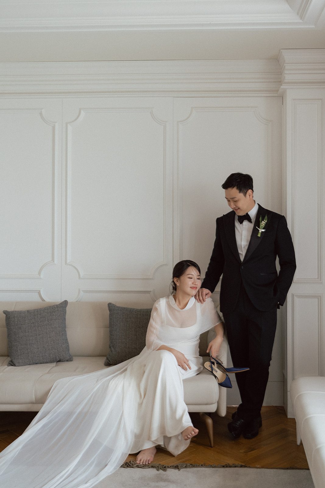 A bride in a long white gown sits on a couch holding a bouquet, while a groom in a black tuxedo stands beside her, both indoors against a white paneled wall—perfect inspiration for couples choosing wedding venues in Korea.