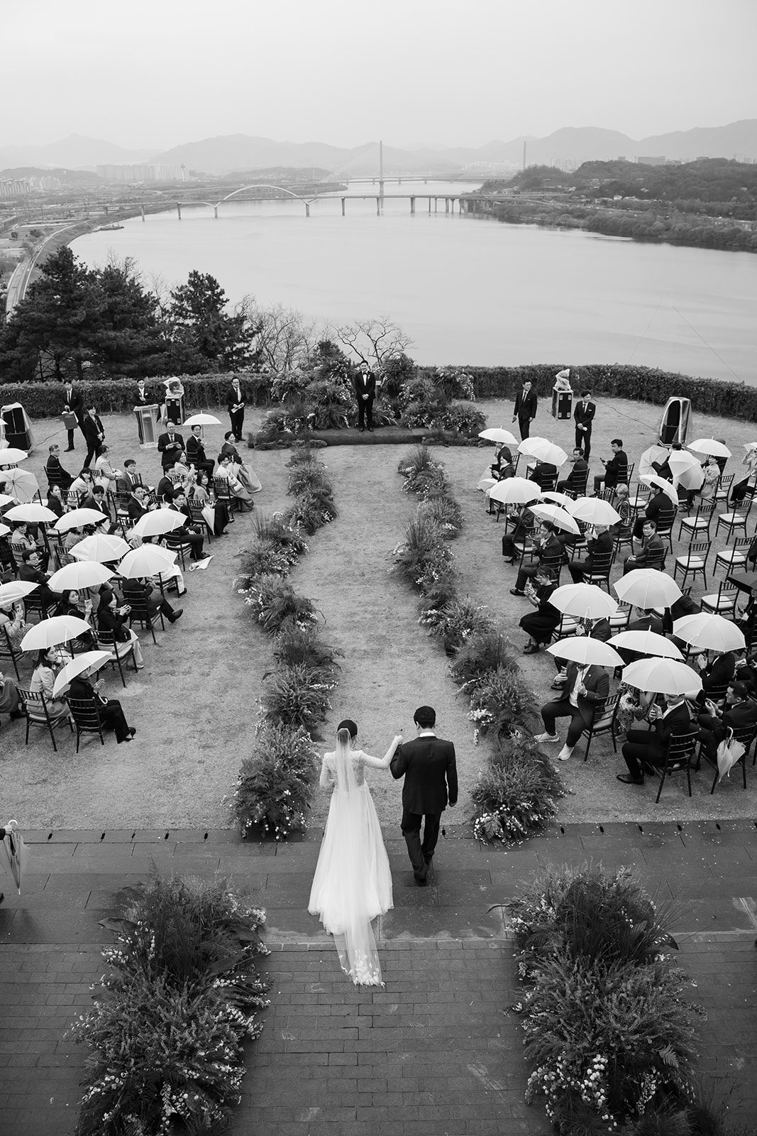 A bride and groom walk down an outdoor aisle toward a wedding ceremony with guests holding umbrellas, overlooking a river and distant mountains—a perfect scene for those choosing wedding venues in Korea.