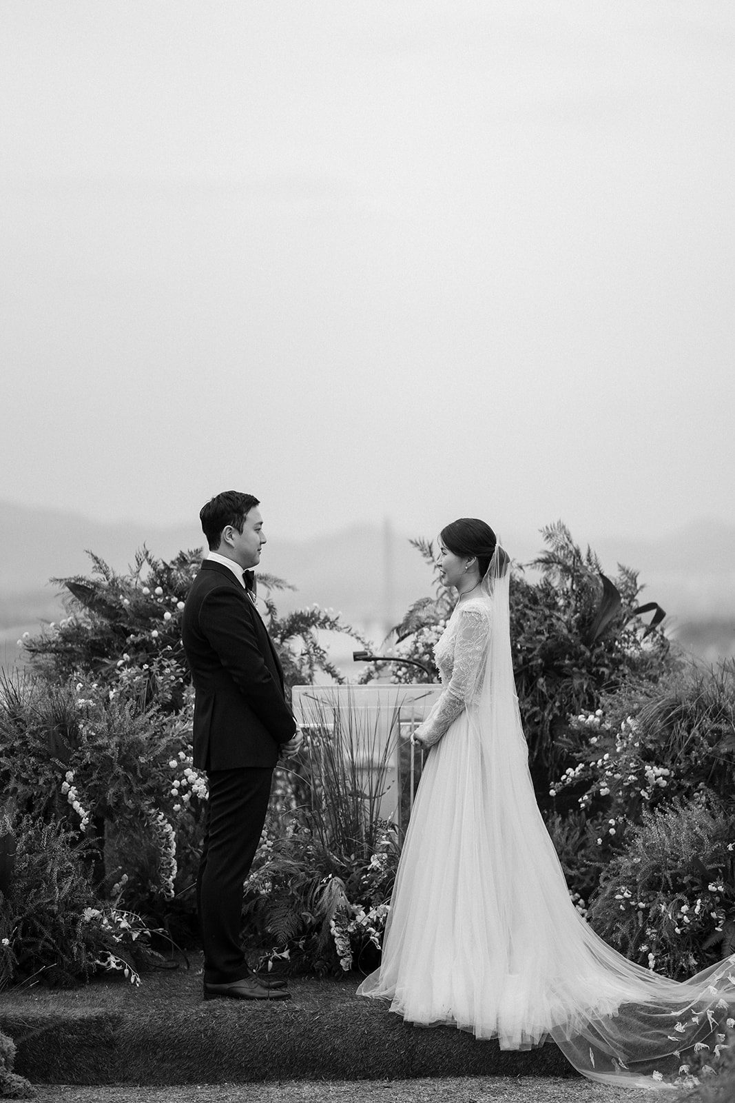 A bride and groom stand facing each other outdoors, surrounded by greenery and flowers, during a wedding ceremony—an inspiring scene for those choosing wedding venues in Korea.