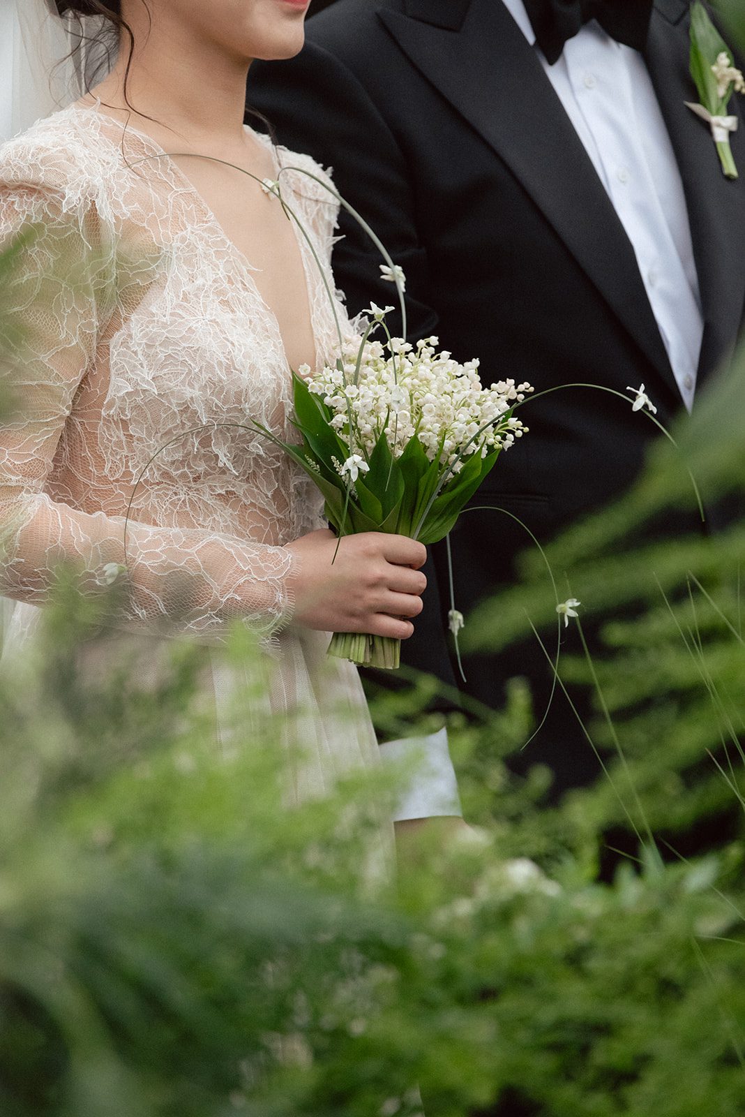 A bride in a lace wedding dress holds a bouquet of lilies of the valley, standing beside her groom in a black tuxedo. Greenery surrounds them—a romantic scene for couples choosing wedding venues in Korea.