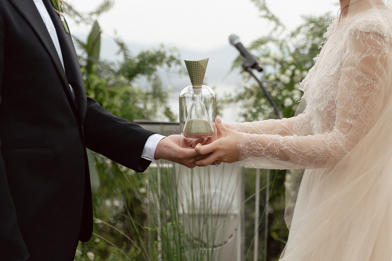 Two people in formal attire hold a glass hourglass together during an outdoor ceremony, surrounded by greenery and a microphone stand—capturing the elegance often considered when choosing wedding venues in Korea.