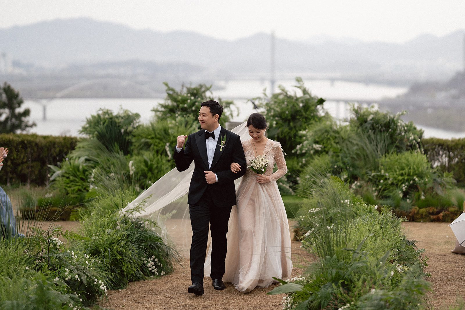 A bride and groom walk arm in arm outdoors, surrounded by greenery, with a river and mountains visible in the background—a scene that captures the charm of choosing wedding venues in Korea.