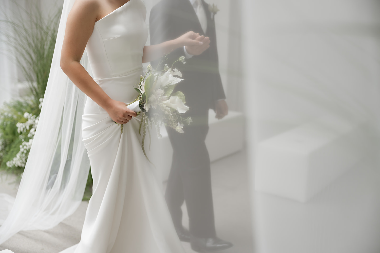 A bride in Korea in a white dress holding a bouquet walks with a man in a dark suit, partially obscured by a sheer veil or curtain on her wedding day. 