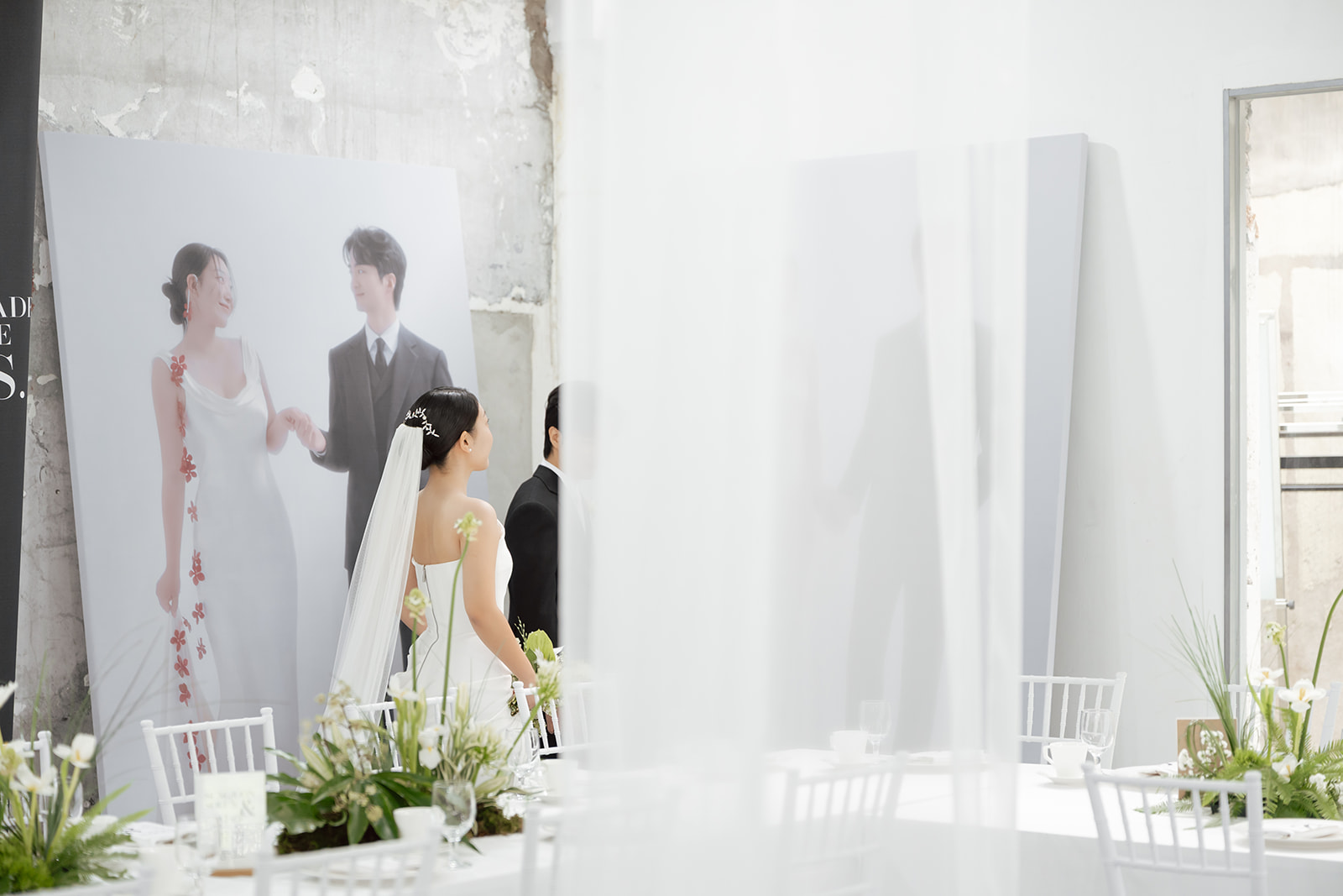 A bride and groom in Korea stand indoors beside a large wedding day portrait of themselves; the setting features white decor and green floral arrangements.