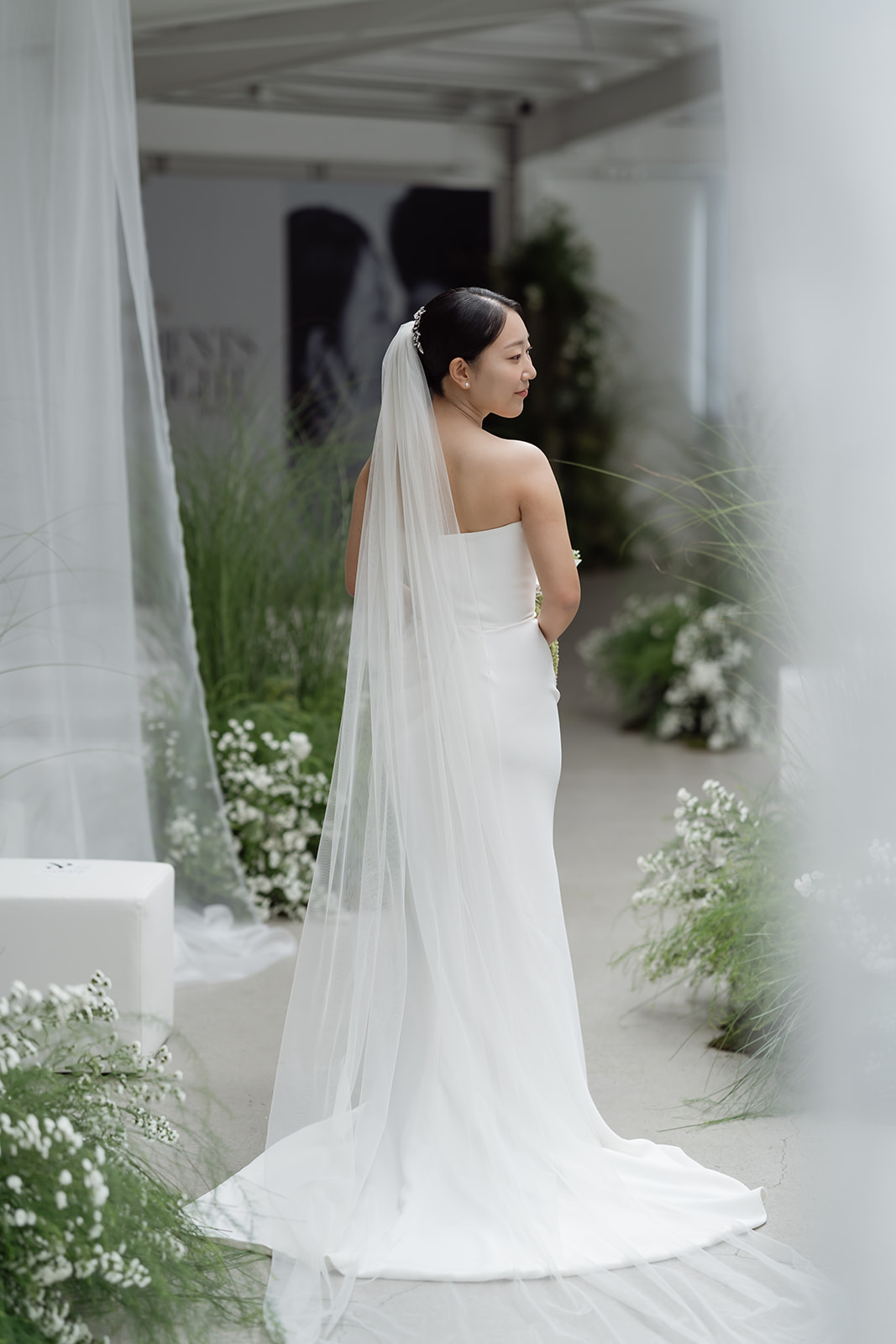A bride in Korea in a white, strapless gown with a long veil stands among greenery and white flowers, facing away from the camera on her wedding day. 
