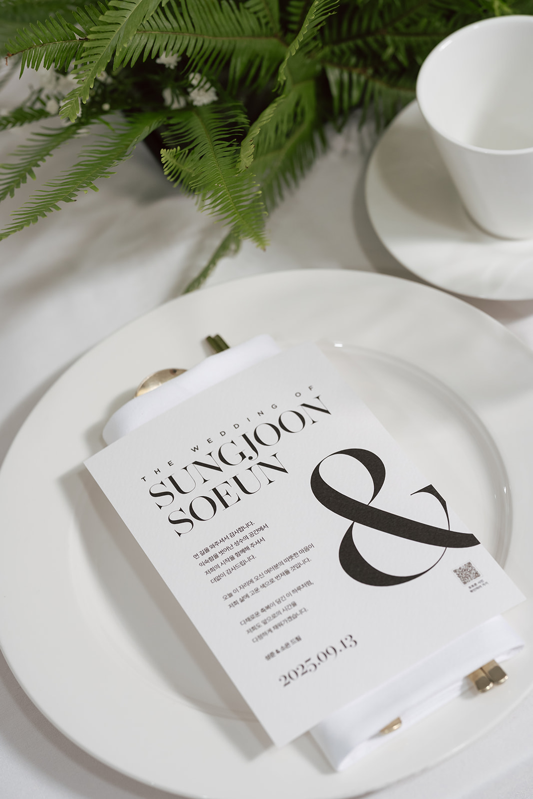 A wedding invitation card for Sungjoon and Soeun placed on a white plate, with a cup and greenery in the background.