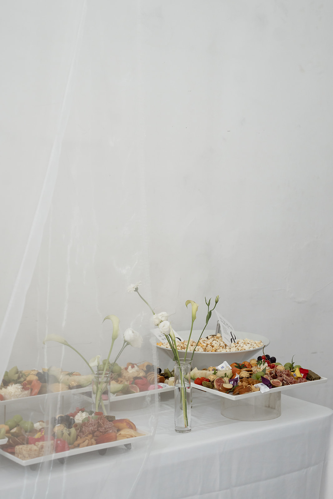 A table set with assorted platters of food and a vase of white flowers, partially obscured by a sheer white curtain against a plain white wall.