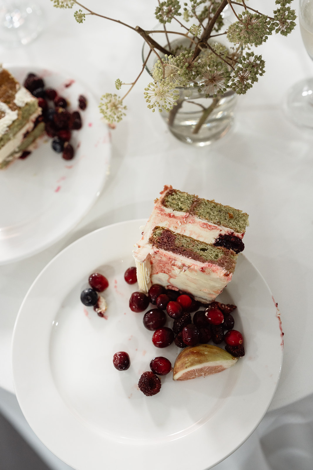 A slice of layered cake with berries and a fig wedge on a white plate, next to a partially eaten piece of cake and a small vase with flowers on a white tablecloth.
