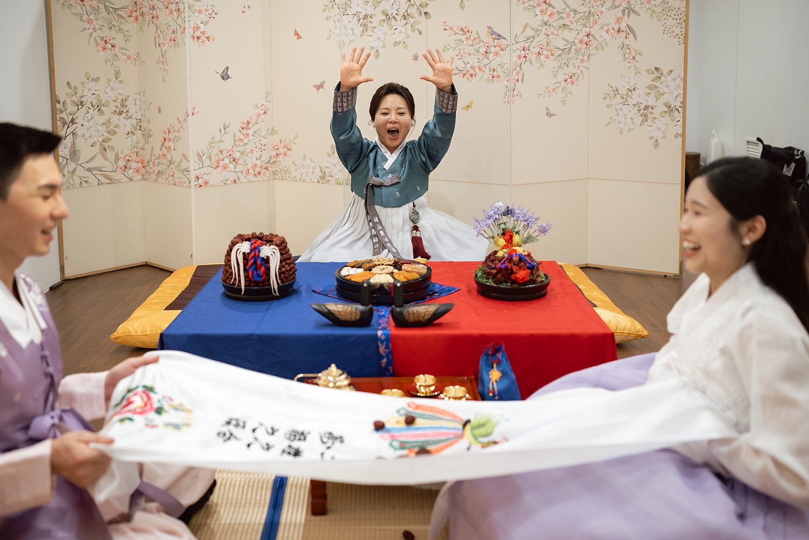 Three people in traditional Korean clothing celebrate a special occasion with food, decorations, and embroidered cloth in a decorated room. One person stands with raised arms, smiling.