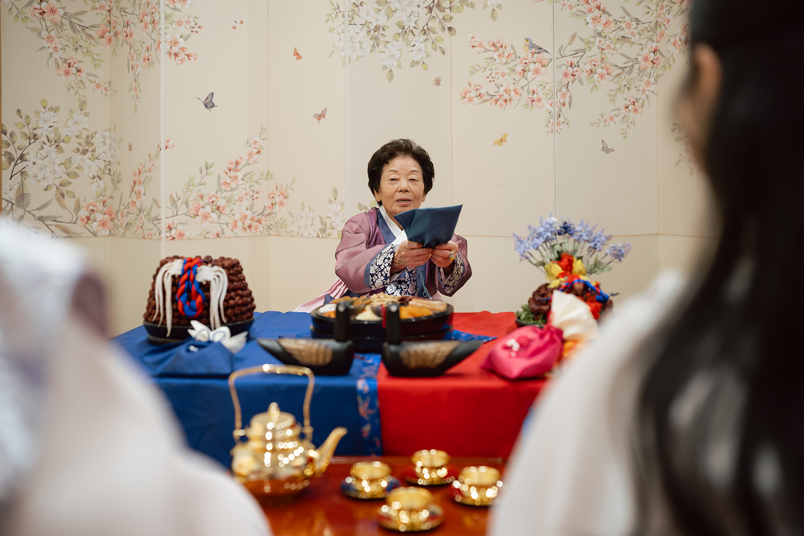 An elderly woman in traditional Korean attire holds a blue envelope across a decorated table with food, teaware, and wrapped gifts, with two people in hanbok in the foreground at a korean wedding celebration.