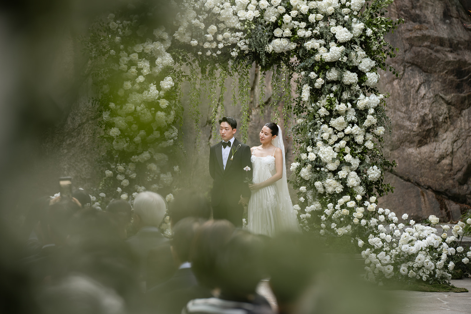 A bride and groom stand together under a large floral arch during an outdoor wedding ceremony, with guests seated in the foreground.