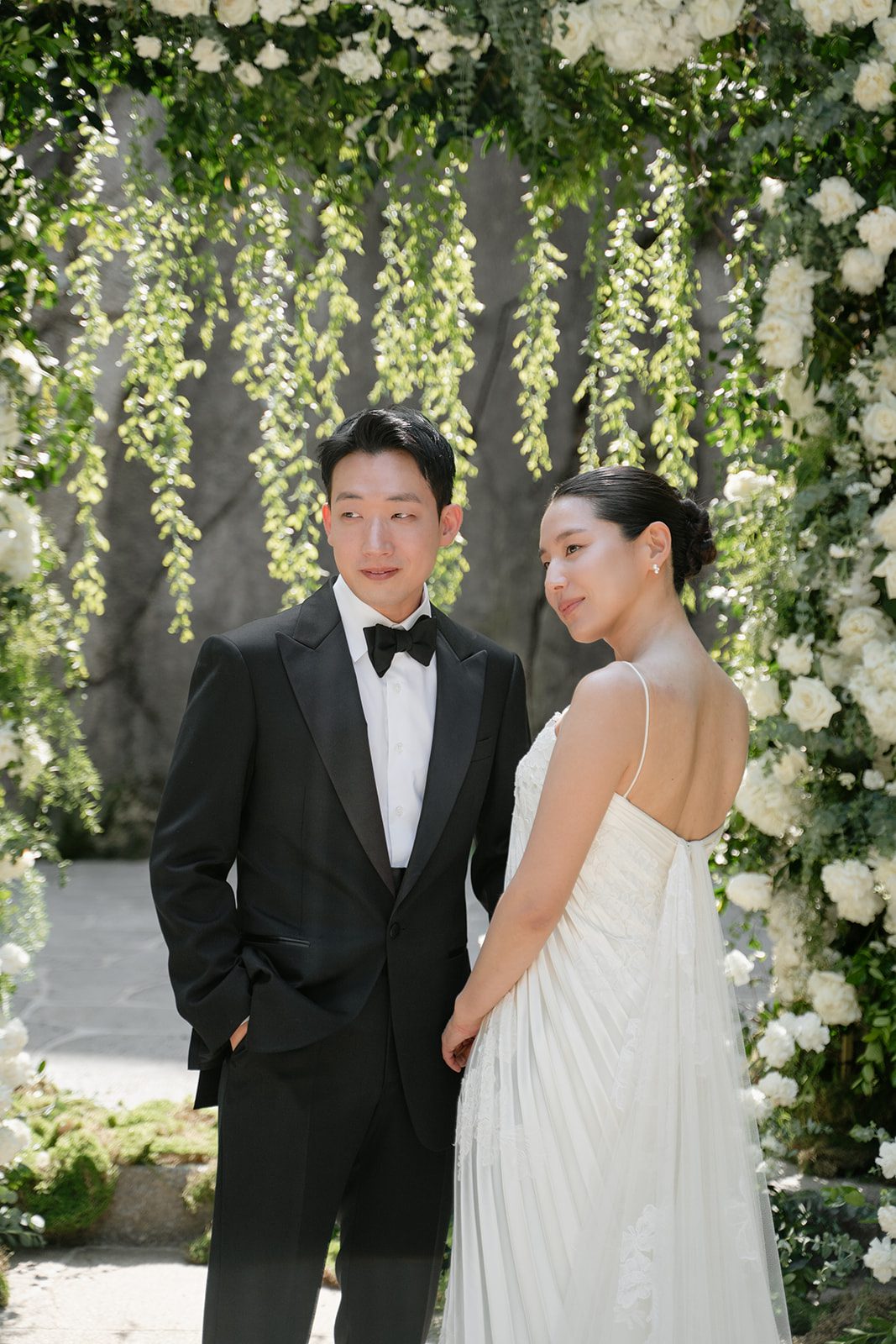 A couple stands together in formal wedding attire under an arch decorated with white flowers and greenery, capturing the elegance often sought when choosing wedding venues in Korea.