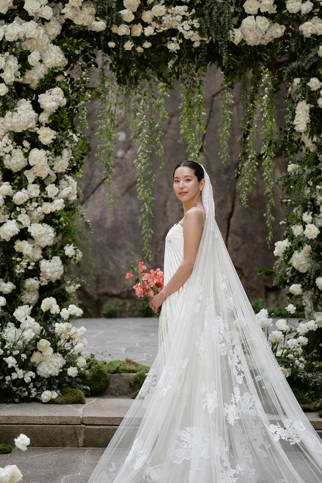 A bride in a white Danielle Frankel gown and veil stands holding pink flowers under a floral arch decorated with white roses and greenery, capturing the charm often found when choosing wedding venues in Korea.