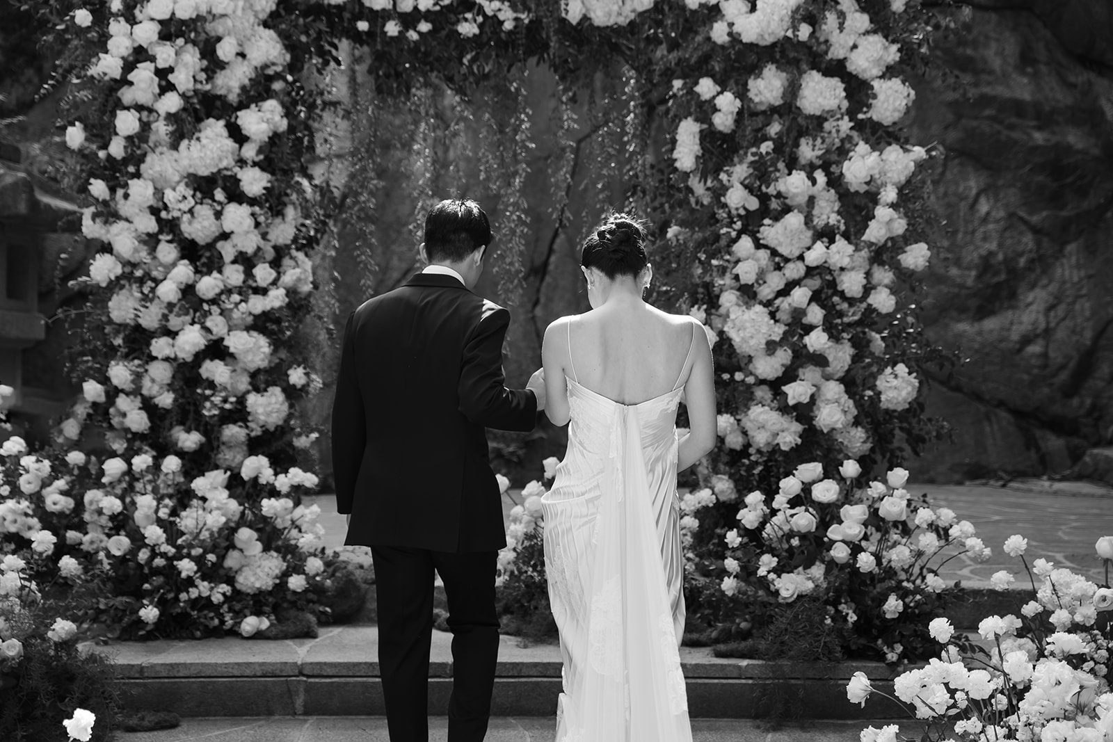 A couple dressed formally walks side by side toward a flower-covered archway in an outdoor setting, reflecting the elegance of choosing wedding venues in Korea.