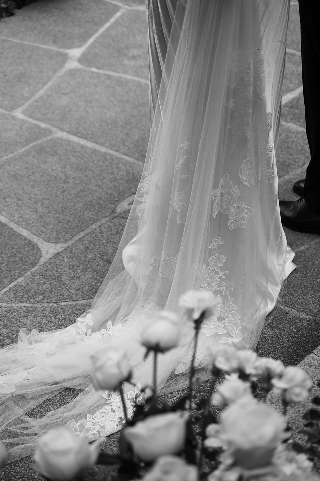 Black and white photo of a bride in a lace wedding dress with a long train standing next to a groom, with flowers in the foreground—perfect inspiration for couples choosing wedding venues in Korea.