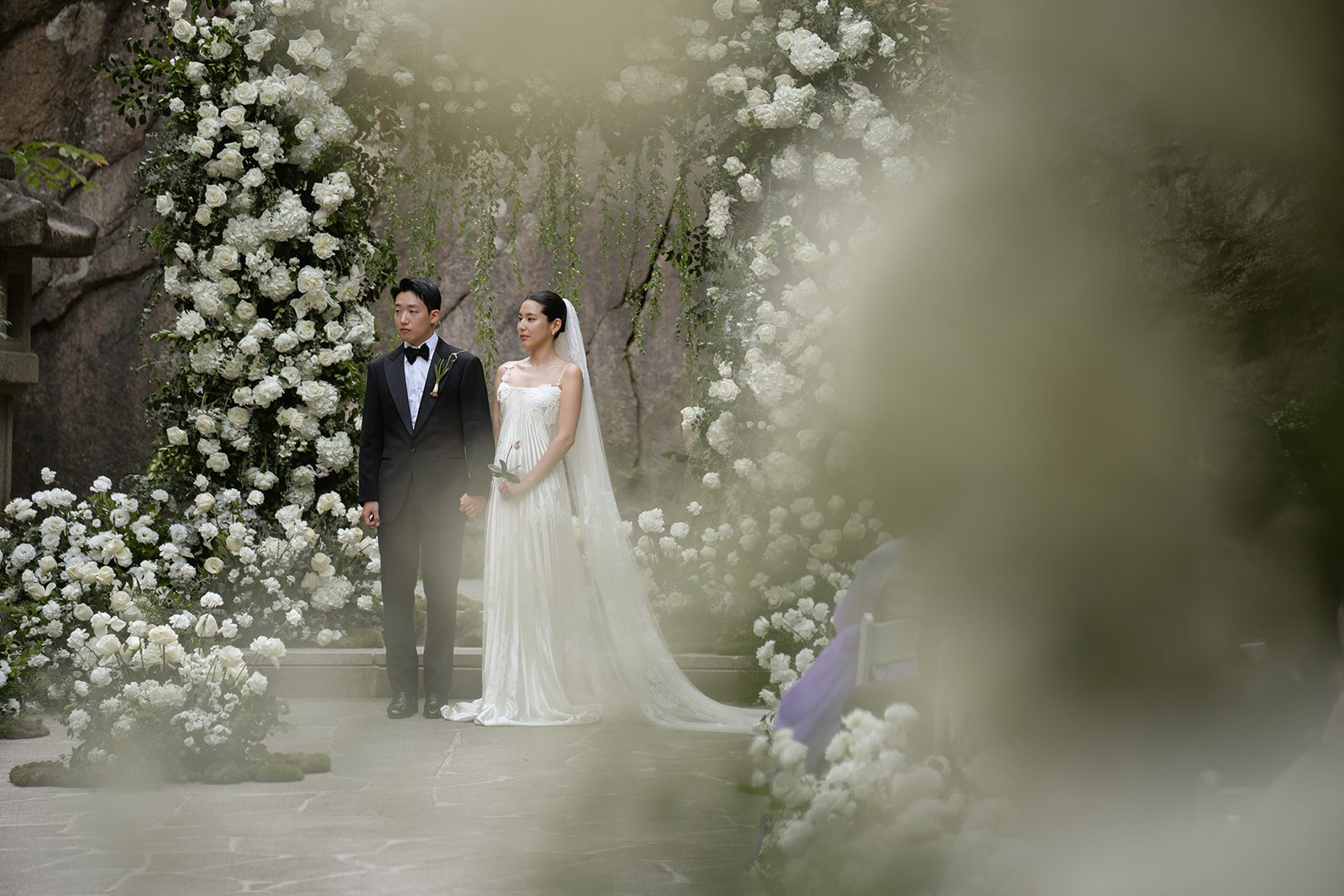 A bride and groom stand together at an outdoor wedding ceremony, surrounded by white flowers and greenery—an elegant scene that inspires couples choosing wedding venues in Korea.