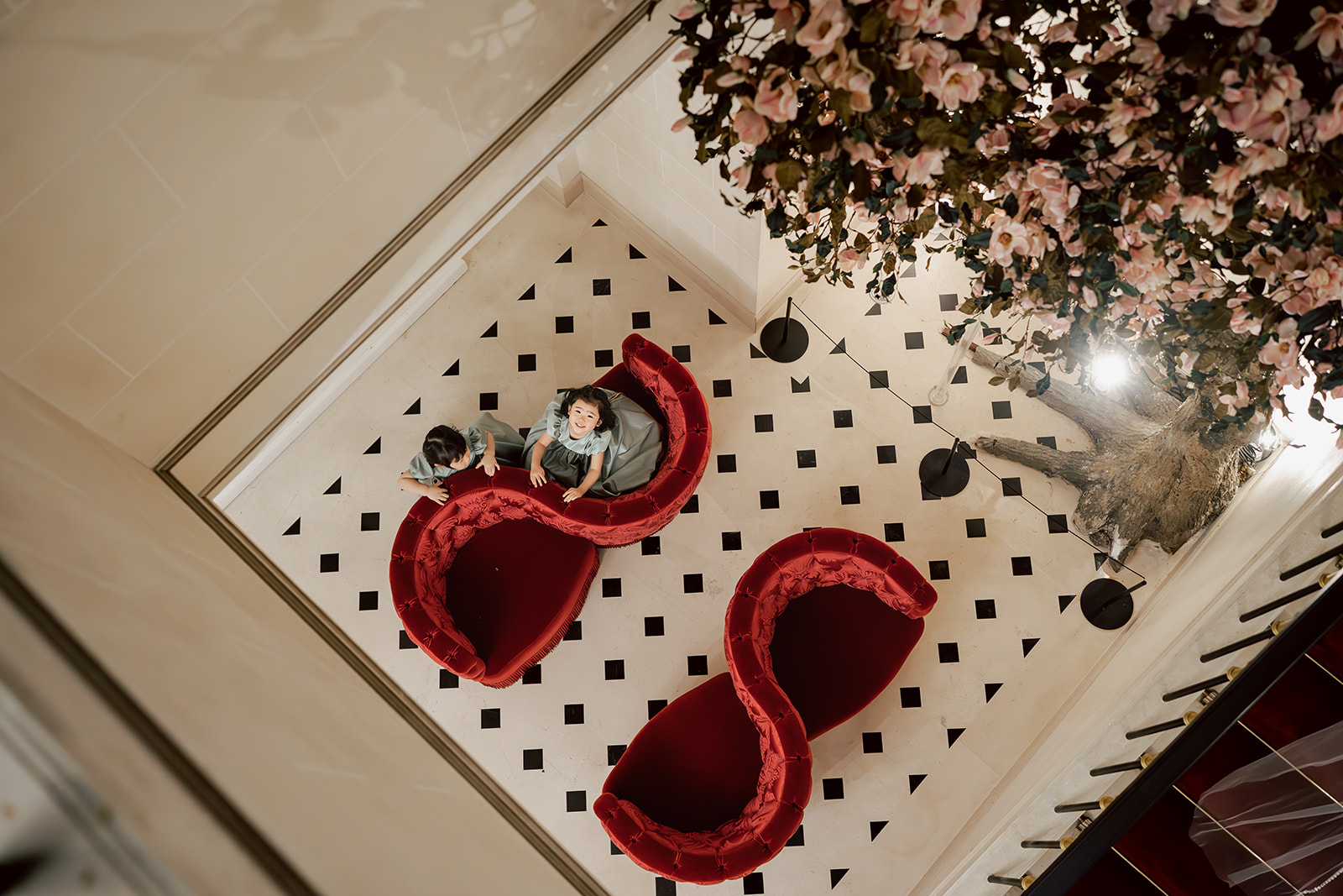 Two people sit on red, S-shaped couches in a tiled room with a black and white floor, seen from above beside a large floral arrangement.