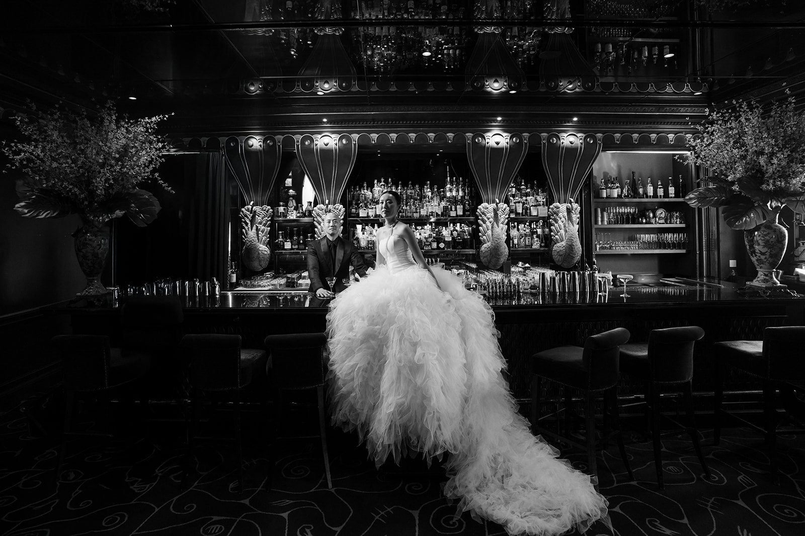 A bride in a voluminous wedding gown stands at a bar beside a bartender in an elegant, dimly lit setting.