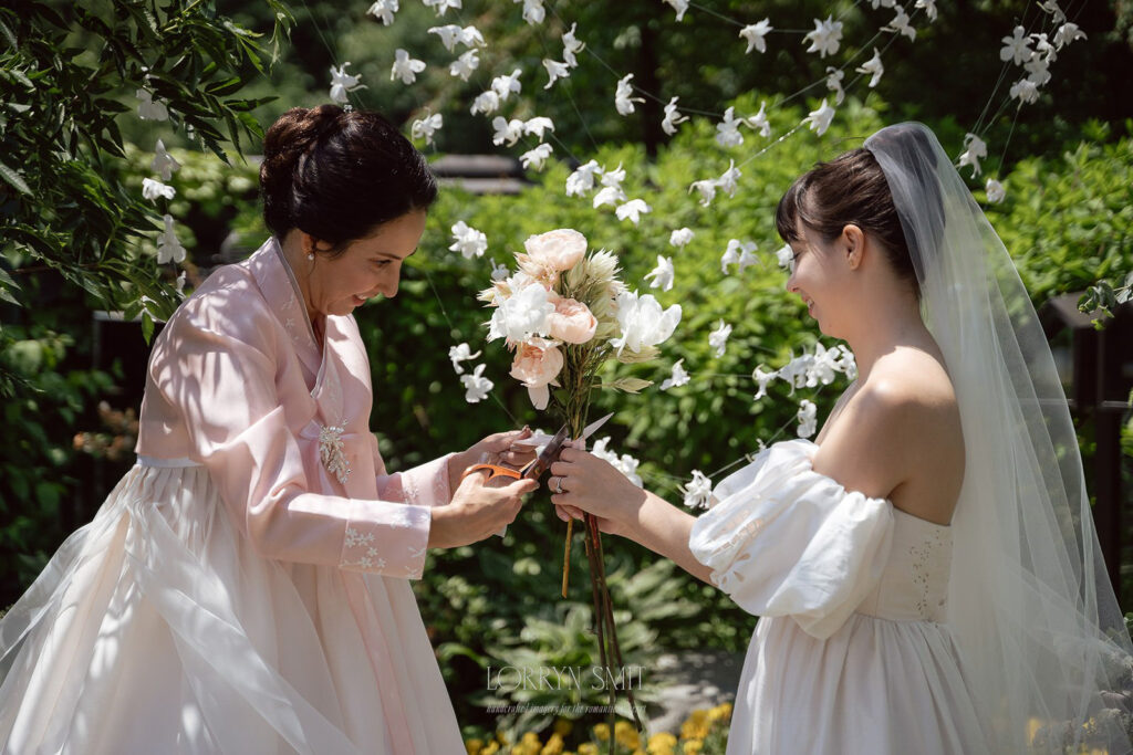 Two women stand outdoors at a Samcheonggak wedding; one in a pink hanbok adjusts flowers held by the other in a white dress and veil. White flowers are strung in the background.