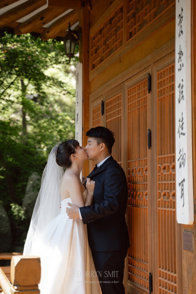 A bride and groom in formal attire share a kiss in front of a hanok with vertical signage and decorative doors.