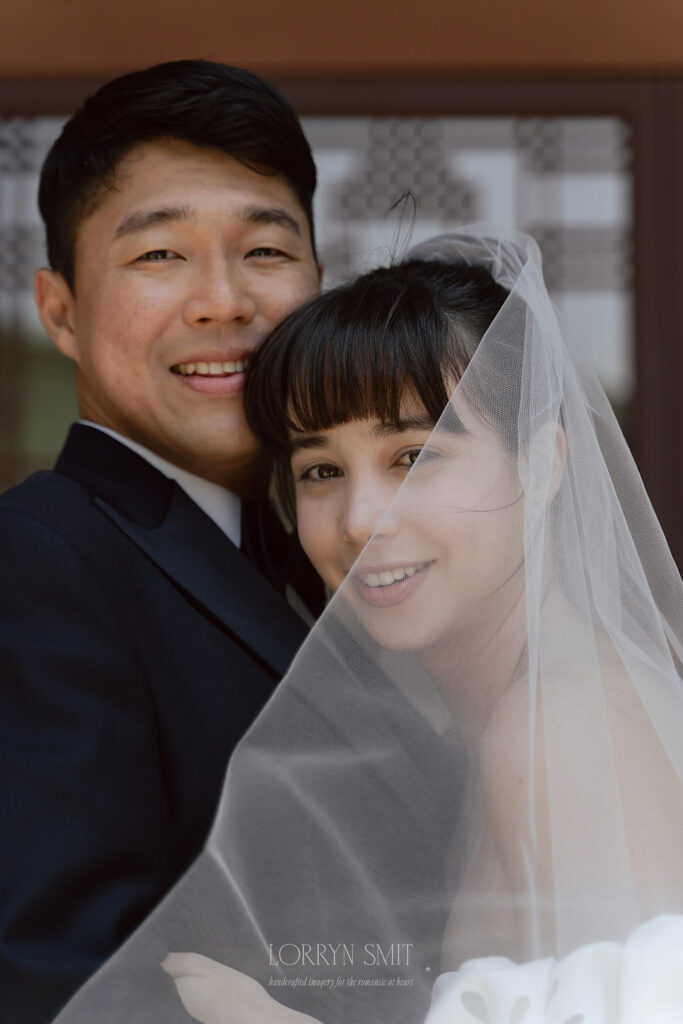 A couple in wedding attire smiles at the camera during their Samcheonggak wedding; the bride's veil partially covers both their faces.