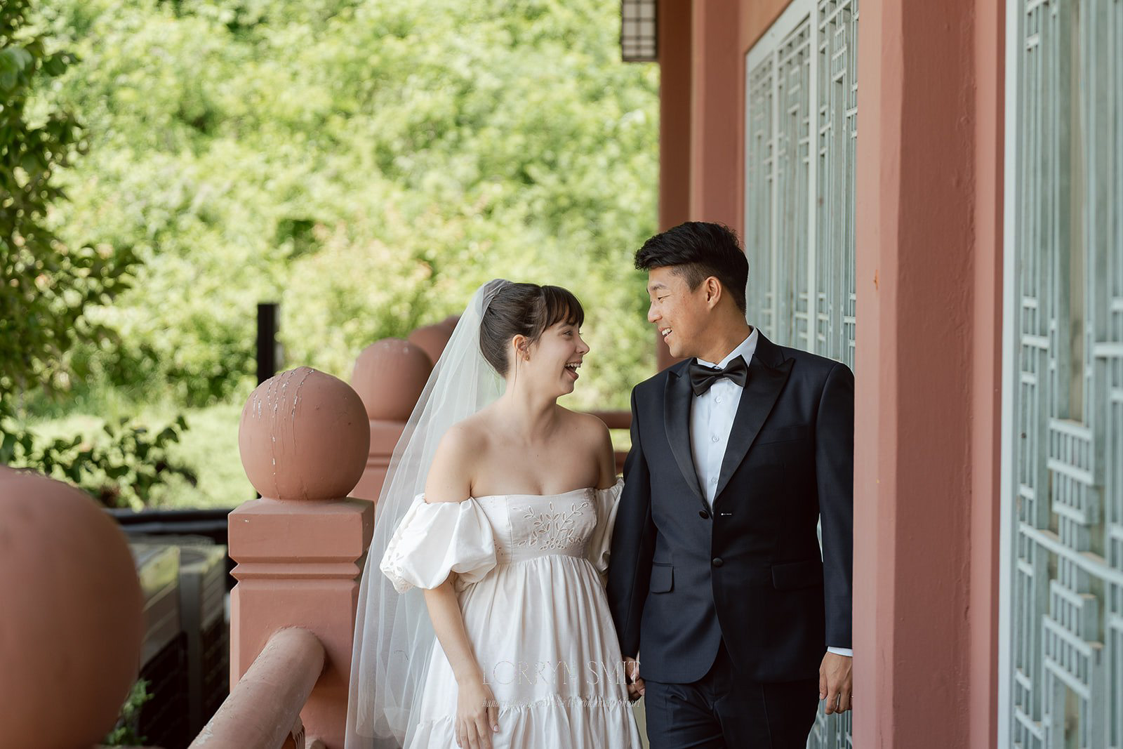 A bride in a white dress and veil holds hands with a groom in a black tuxedo as they smile at each other on an outdoor balcony during their elegant Samcheonggak wedding.