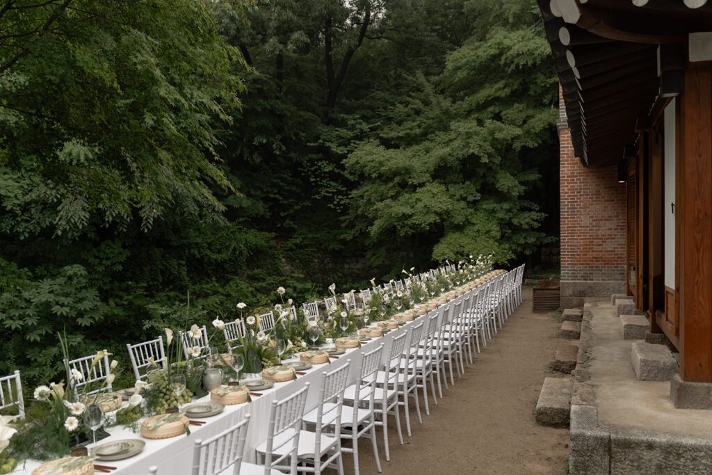 A long outdoor dining table with white chairs is set with plates and decorated with green foliage and white flowers, perfect for those choosing wedding venues in Korea beside a traditional building and lush trees.