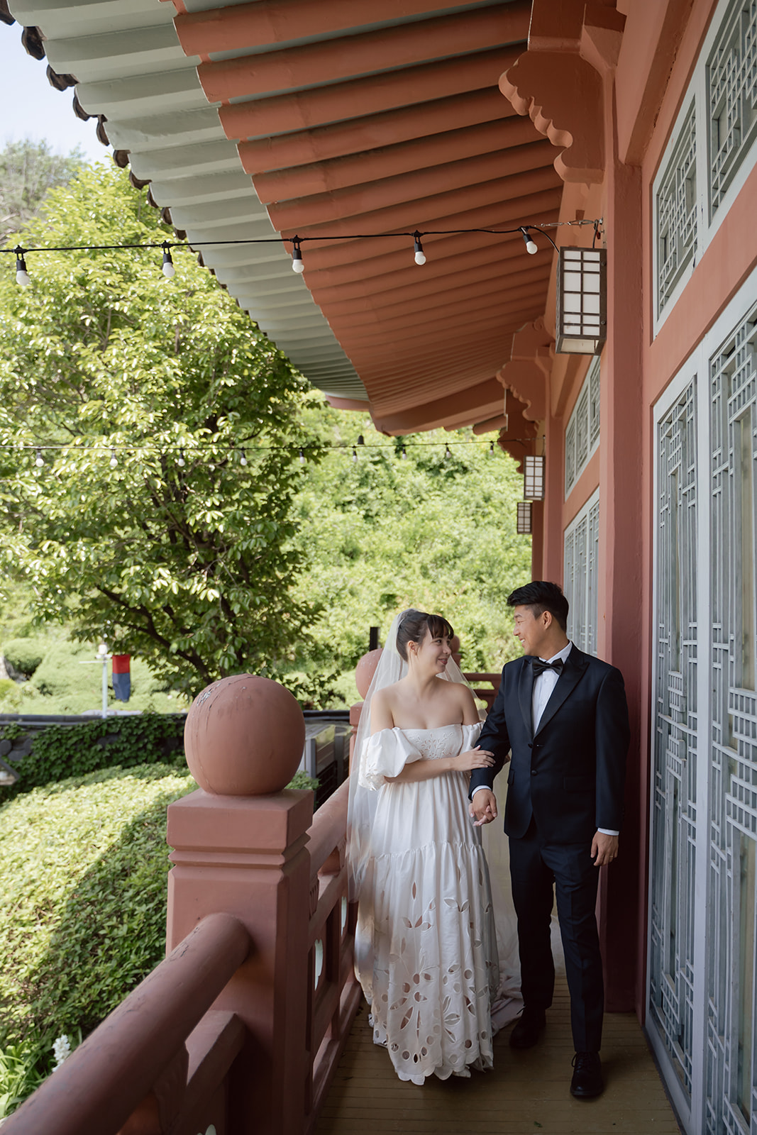 A bride and groom in Korea walk together on a balcony with traditional architecture, surrounded by greenery during their wedding day.