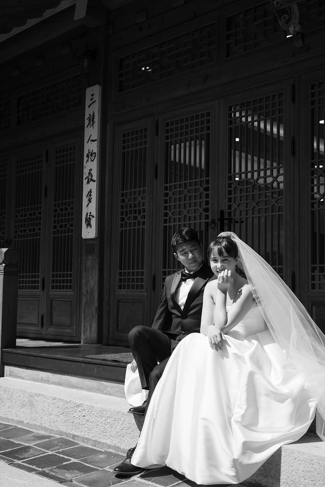A bride and groom in Korea in formal wedding attire sit on steps outside a traditional building with wooden doors and a vertical sign featuring Asian script, capturing the elegance highlighted in any wedding day. 