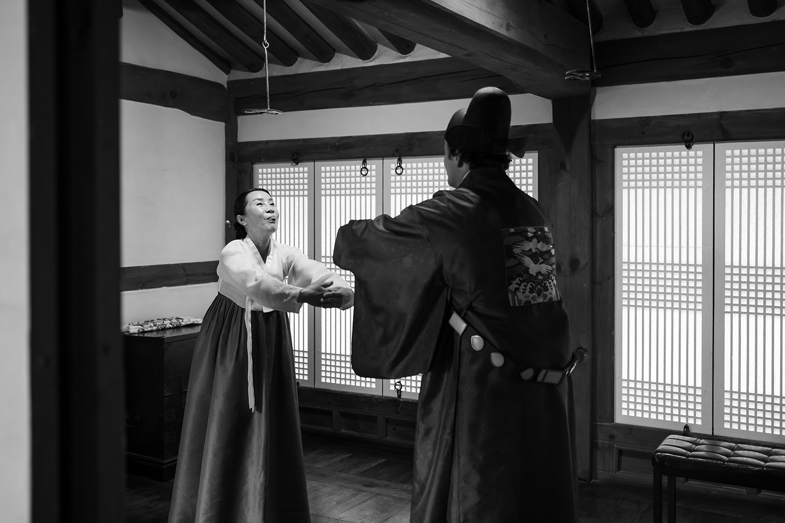 Two people in traditional Korean hanbok stand facing each other inside a wooden room, one smiling and extending their hands forward—a scene reminiscent of a korean wedding.